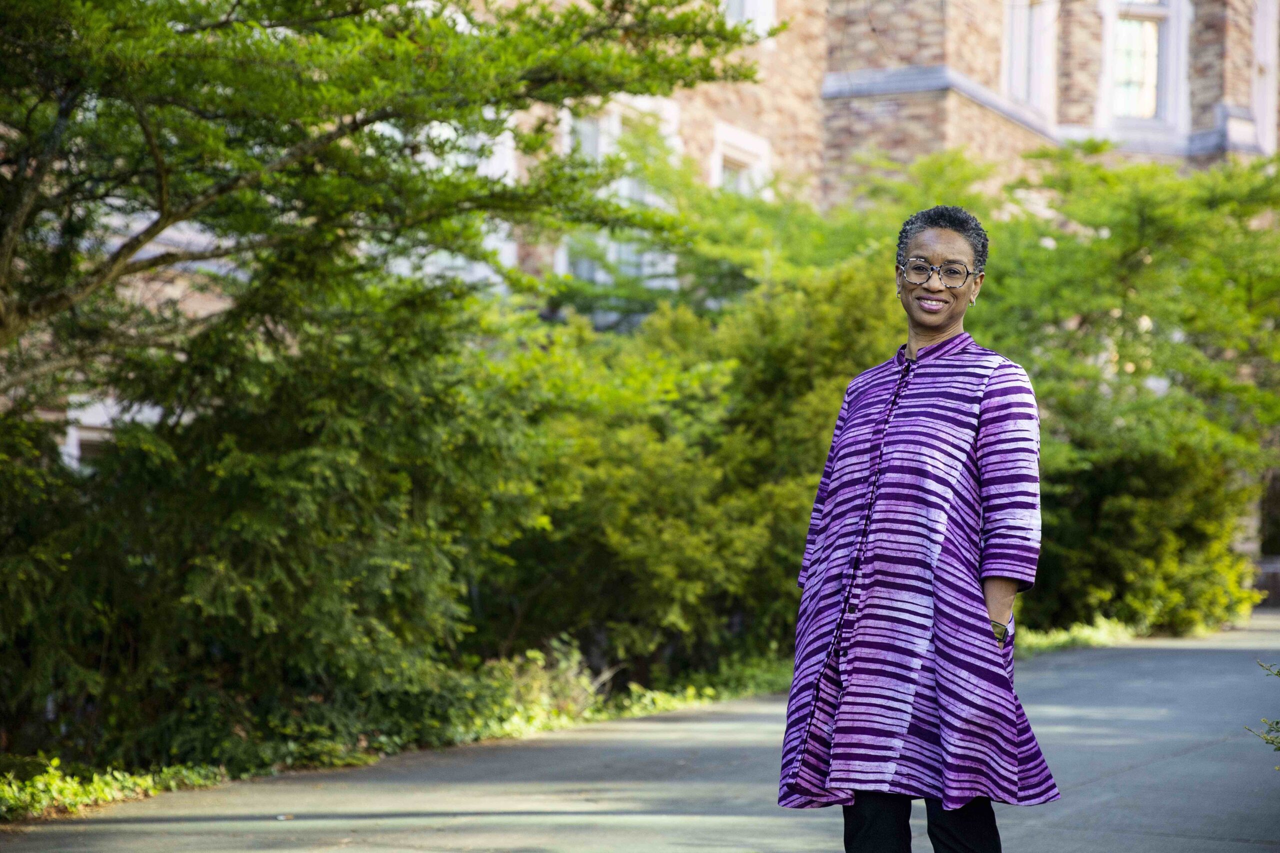 Stephanie Smallwood Honors Director standing in front of green trees and bushes outside of mary gates hall during a sunny day, wearing a striped purple coat
