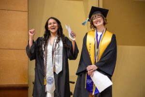two Honors students in graduation gowns one with arms in the air cheering, other wearing cap and smiling